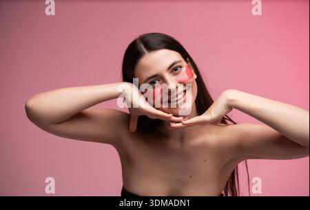 Cheerful young woman with pink under eye patches framing face with hands on pastel background, playful skincare beauty portrait Stock Photo