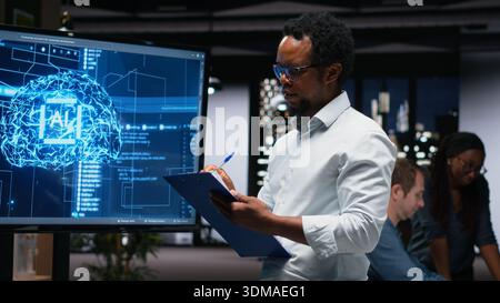 Manager in office writing on notepad, taking notes of artificial intelligence system bugs to fix. Supervisor looking at large screen, documenting neural network errors during troubleshooting Stock Photo