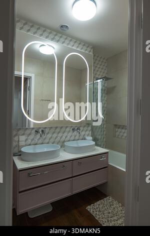 Contemporary bathroom interior shows twin oval vessel sinks, LED halo mirrors, a bathtub with glass screen and rainfall shower, beige tiles, and warm Stock Photo