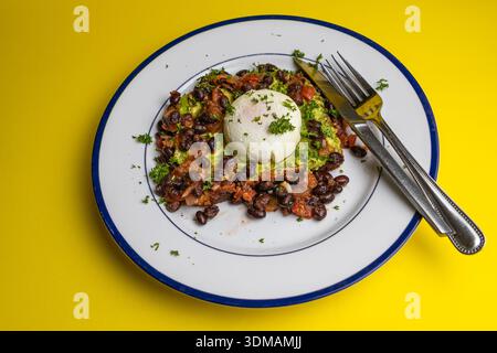 poached egg on whole wheat toast spread with avocado  and served with black beans and tomato salsa on a blue rimmed plate. On a yellow background Stock Photo