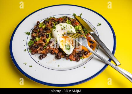 poached egg on whole wheat toast spread with avocado  and served with black beans and tomato salsa on a blue rimmed plate. On a yellow background Stock Photo