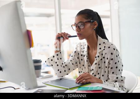 African American woman wearing polka-dot blouse sitting at desk holding stylus on graphics tablet. Designer, illustrator, professional, workspace, mod Stock Photo