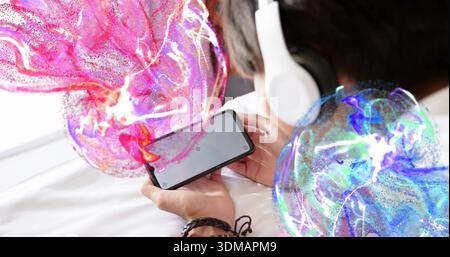 Holding smartphone in both hands, lying teen boy on bed wearing white headset, holographic spheres Stock Photo