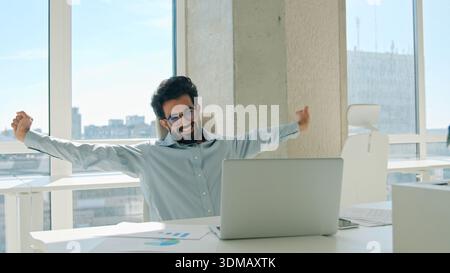 Happy Arabian Indian business man office employee relax stretching after laptop work tired businessman male entrepreneur taking break relief tension Stock Photo