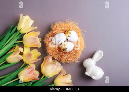 Easter eggs in decorative nest with yellow tulips and white ceramic bunny on pastel background. Top view, flat lay Stock Photo