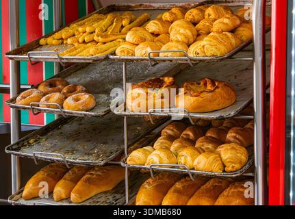 A variety of bread and pastries are neatly arranged on metal trays in a bakery display. The golden hues of the baked items shine under bright lights. Stock Photo