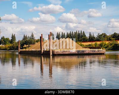 A rusty flat-deck barge carrying large piles of brown earth sits on the calm waters of the Sacramento River. Stock Photo