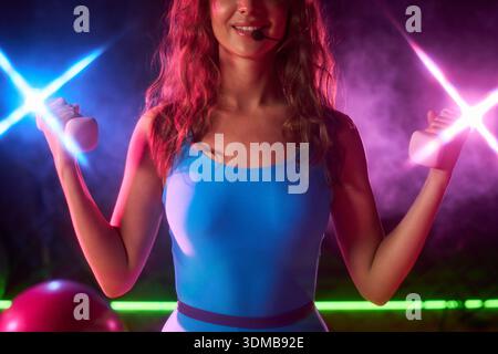 Young adult Caucasian woman leading fitness class, holding dumbbells in both hands, wearing headset microphone, smiling while exercising in gym with colorful lights in background Stock Photo