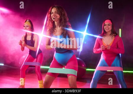Three young adult Caucasian women exercising with resistance bands, leading group fitness class on stage, smiling and performing squats, wearing retro workout headbands and leotards Stock Photo