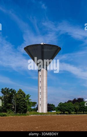 Modern concrete water tower rising above a rural field and green trees under a blue sky with wispy clouds. Stock Photo