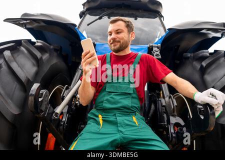 Male farmer using his smartphone while leaning on tractor outdoors. Stock Photo