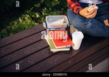 Close up view of student that is eating sandwich, sitting with notepads, an apple and cup of drink. Stock Photo