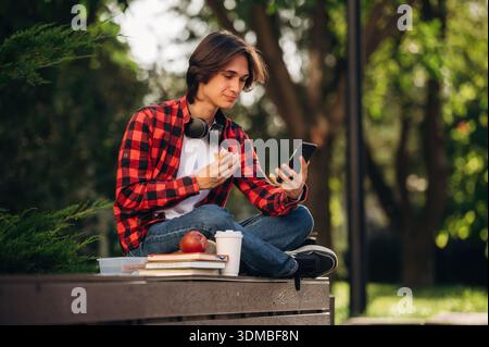 Eating, sitting and using smartphone. Male student is in the public park outdoors. Stock Photo
