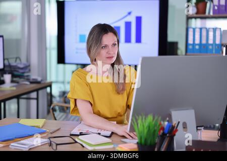 Woman in yellow shirt focuses on computer screen while analyzing business growth in a modern office setting Stock Photo
