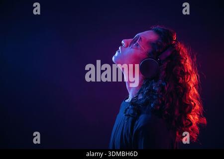 Long hair, headphones, side view. Guy is indoors at night with neon lighting. Stock Photo
