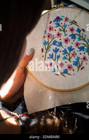 Young woman holding embroidery hoop with colorful floral handmade design in warm sunlight. Close-up of needlework process and creative hobby concept. Stock Photo
