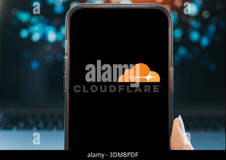 Smartphone displaying the Cloudflare logo on a black screen, held in hand against a blurred technology background. Concept image representing cloud co Stock Photo