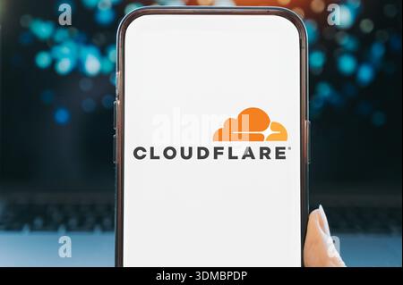 Smartphone displaying the Cloudflare logo on a white screen, held in hand against a blurred background. Concept of cloud computing, web security, cont Stock Photo