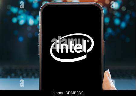 Smartphone displaying the Intel logo on a black screen, held in hand against a blurred technology background. Concept image representing semiconductor Stock Photo