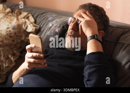 Middle aged man lying on a sofa at home feeling stressed by digital notifications and smartphone alerts. Concept of information overload, mental fatig Stock Photo