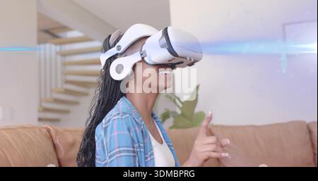 Pointing woman wearing white VR headset, blue plaid shirt, sitting on tan couch in living room Stock Photo