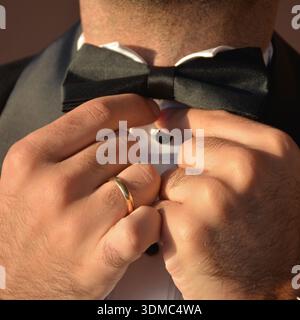 Groom Adjusting Black Bow Tie Close Up Preparation For Wedding Ceremony. Wearing a black tuxedo suit and a white shirt with black buttons. Detail of h Stock Photo