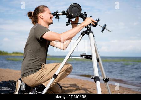 young man prepares his astronomical telescope for use on a sandy beach. astronomer is focused on adjusting equipment under clear blue skies, ready to observe solar activity as part of astronomy hobby Stock Photo