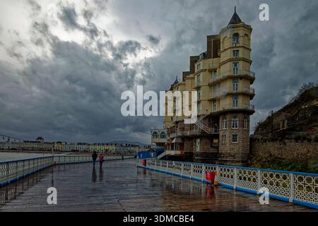 A couple (man and woman) walk along a pier toward a seafront with a towering hotel in the rain as storm clouds roll in over Llandudno, North Wales. Stock Photo