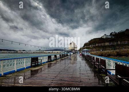 Storm clouds billow over a coastal resort with a deserted rain soaked pier walkway leading to a seafront hotel, Llandudno, North Wales. Stock Photo