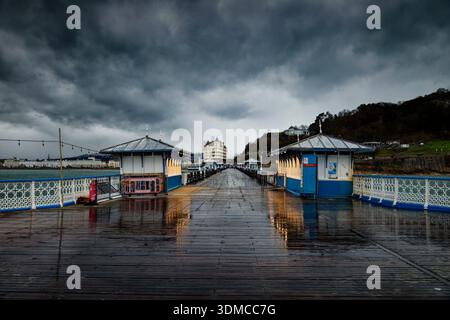 Dramatic billowing storm clouds roll in over a wet and deserted pier with closed kiosks and a seafront hotel at Llandudno, North Wales. Stock Photo