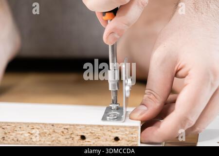 A focused close-up of hands using a screwdriver to fasten a metal bracket onto a wooden board, emphasizing practical DIY skills, home improvement, and Stock Photo