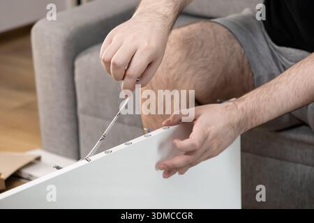 Close-up of a person's hands diligently working on a DIY project, assembling new white flat-pack furniture with a screwdriver in a modern home interio Stock Photo