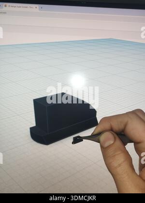 Detail of a hand of a craftsman mechanic holding a small piece with tweezers in front of the computer screen that shows the original project designed Stock Photo