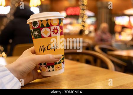 Poznan, Poland - November 19, 2025: Hand holding a McCafe disposable coffee cup with a festive pattern, enjoying a comfortable moment inside a blurred Stock Photo