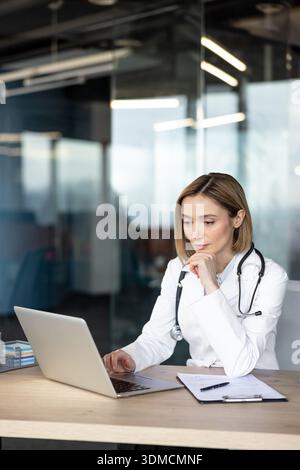 Female doctor in white coat and stethoscope using a laptop at a wooden desk in a contemporary healthcare setting, thinking deeply while reviewing medical records and patient information Stock Photo