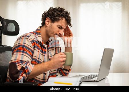 Overworked young businessman having a headache and drinking coffee while working on laptop at his home. Stock Photo