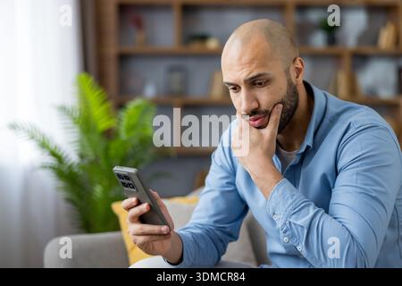 Young adult man sitting on a sofa at home, feeling worried and pensive while looking at his mobile phone, reflecting on content he is seeing on the screen Stock Photo