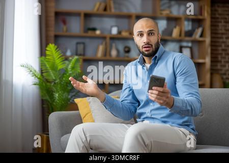 Surprised man sitting on sofa, holding mobile phone, feeling disbelief and confusion while reacting to unexpected shocking news or a surprising message at home Stock Photo