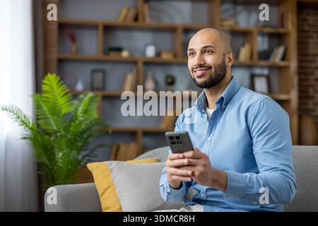 Young man smiling and relaxed on a sofa, casually using his smartphone to browse social media and connect online from his comfortable living room Stock Photo