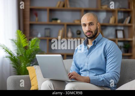 Young man working remotely on a laptop while sitting comfortably on a gray sofa at home, managing his professional tasks in a relaxed yet productive environment Stock Photo