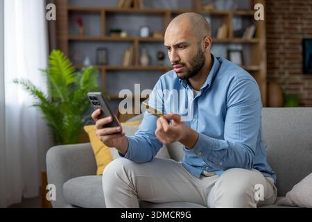 Young man sitting on a sofa at home, holding a smartphone and credit card, looking worried and focused while contemplating an online purchase or checking a transaction, experiencing financial doubt Stock Photo