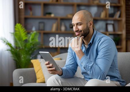 Happy millennial man relaxing on a modern couch, smiling as he reads and browses on a portable tablet, enjoying leisure time, connectivity and entertainment at home Stock Photo