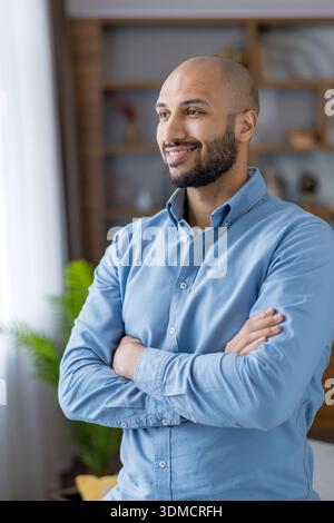 Handsome middle eastern man with beard and bald head smiles confidently, standing indoors with arms crossed in smart-casual attire, conveying success and relaxed professionalism Stock Photo
