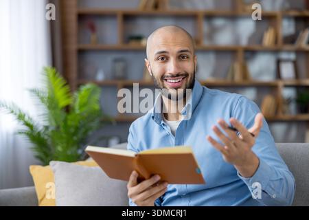 Cheerful male teacher-coach smiles and gestures with a pen while holding a notebook, giving an engaging online lesson or virtual coaching session from his comfortable home office couch Stock Photo
