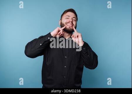 A man stands in front of a blue wall, smiling widely while touching his face. He wears a black button-up shirt and looks cheerful. The setting is info Stock Photo