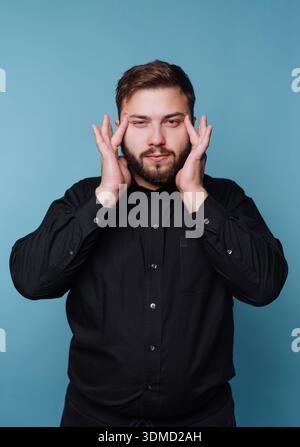 A young man holds his face with both hands while standing against a blue wall. He appears to be deep in thought. The setting looks relaxed and informa Stock Photo