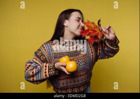 A woman stands holding two oranges in one hand and colorful autumn leaves in the other. She smiles as she enjoys the moment. The background is bright Stock Photo