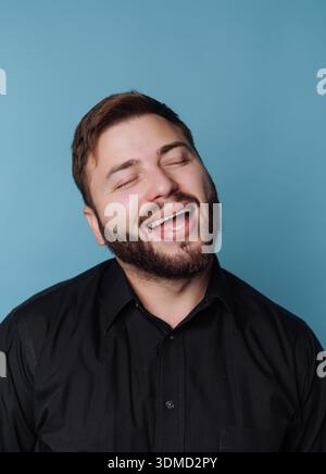 A man with a beard stands indoors with his eyes closed, smiling broadly. He wears a black shirt and shows joy while enjoying a moment. The background Stock Photo