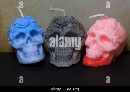 Three artisan skull-shaped candles featuring floral crown detailing, in pastel blue, matte gray, and gradient pink-red tones. Stock Photo