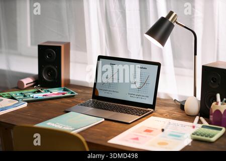 Laptop displaying math graphs on wooden desk surrounded by educational materials, textbooks and stationery suggesting online lesson or remote tutoring session in progress Stock Photo